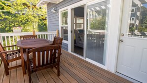 Eine Holzterrasse mit Tisch und Stühlen, eine weiße Tür mit ein Fenstergitter und Blick auf ein Wohngebäude mit Balkonen.