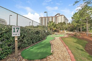 Um campo de minigolfe com área de putting verde, cercado por uma vedação de arame, árvores e um edifício ao fundo.