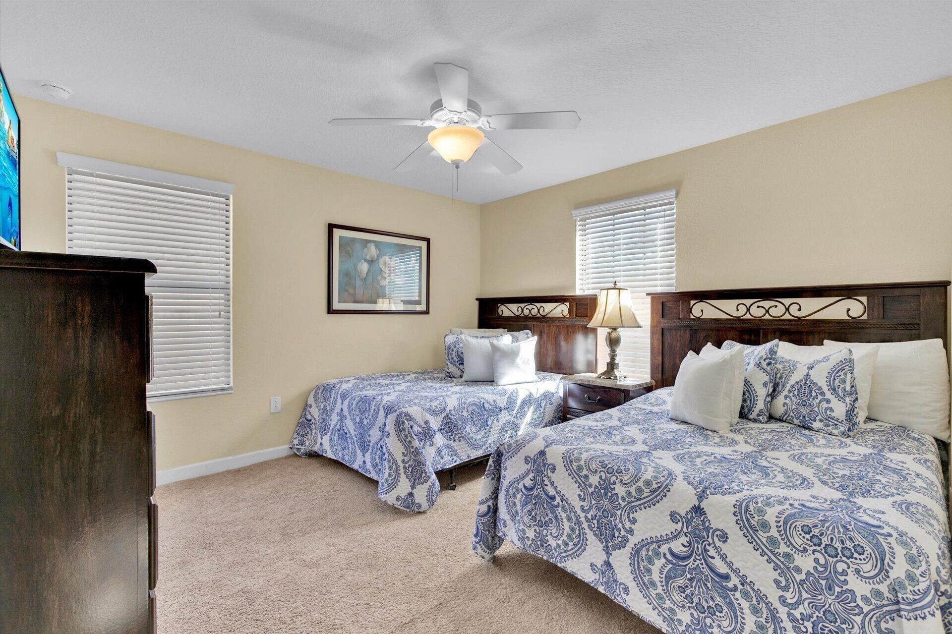 A bedroom with two beds, a ceiling fan, a painting, and a nightstand with a lamp.