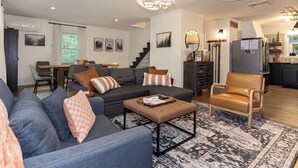 Una sala de estar moderna con un sofá seccional gris, un sillón marrón y una mesa de centro. Se ve una escalera que sube al piso superior, y un área de cocina al fondo.