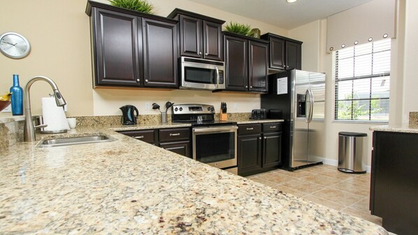 A kitchen with granite countertops, dark cabinetry, stainless steel appliances, and a window with blinds.