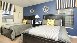 Two single beds with anchor-themed bedding, a ship's wheel accent wall, and a nautical-inspired lamp.
