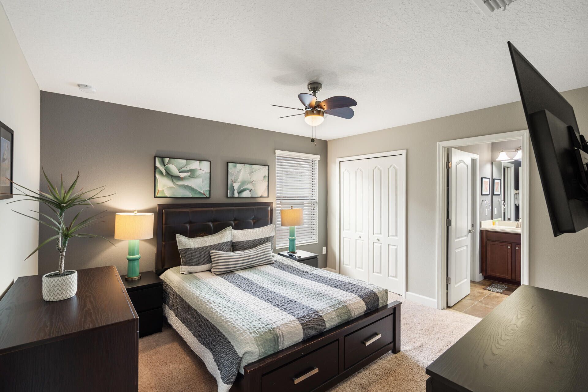 A bedroom with a bed, bedside tables, a ceiling fan, a TV, and a door leading to another room.