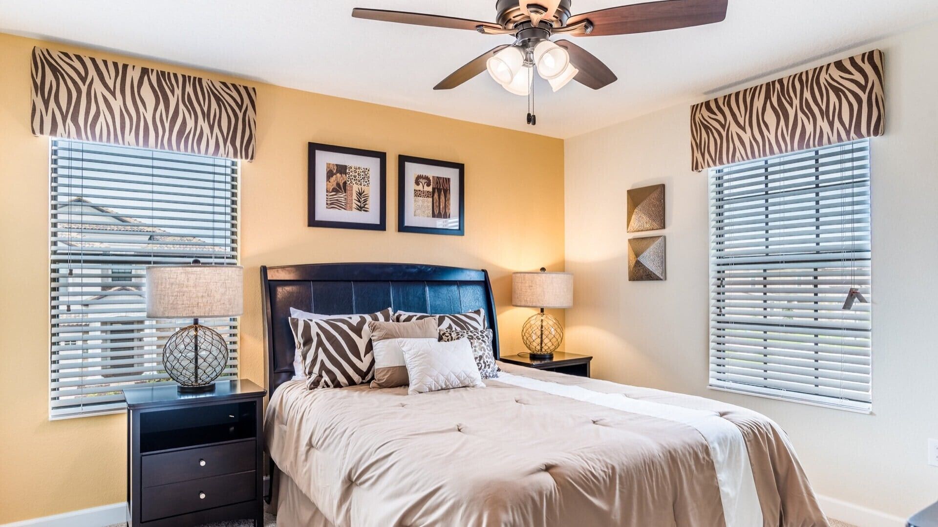 A bedroom with a bed, two nightstands, a ceiling fan, and zebra-patterned window treatments.