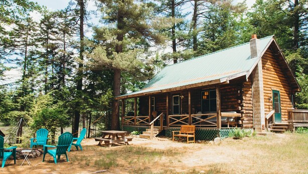 Une cabane rustique en rondins de bois, dotée d’un porche et entourée de grands pins.