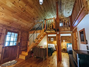 A wooden cabin interior with a staircase leading to a loft bedroom, a dining area, and a kitchen. - Charming Waterfront Log Cabin (Fitzwilliam)