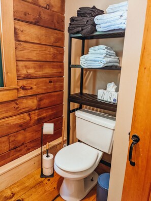 A compact bathroom with a wooden wall, a white toilet, a shelf with folded towels, and a toilet paper holder. - Charming Waterfront Log Cabin (Fitzwilliam)