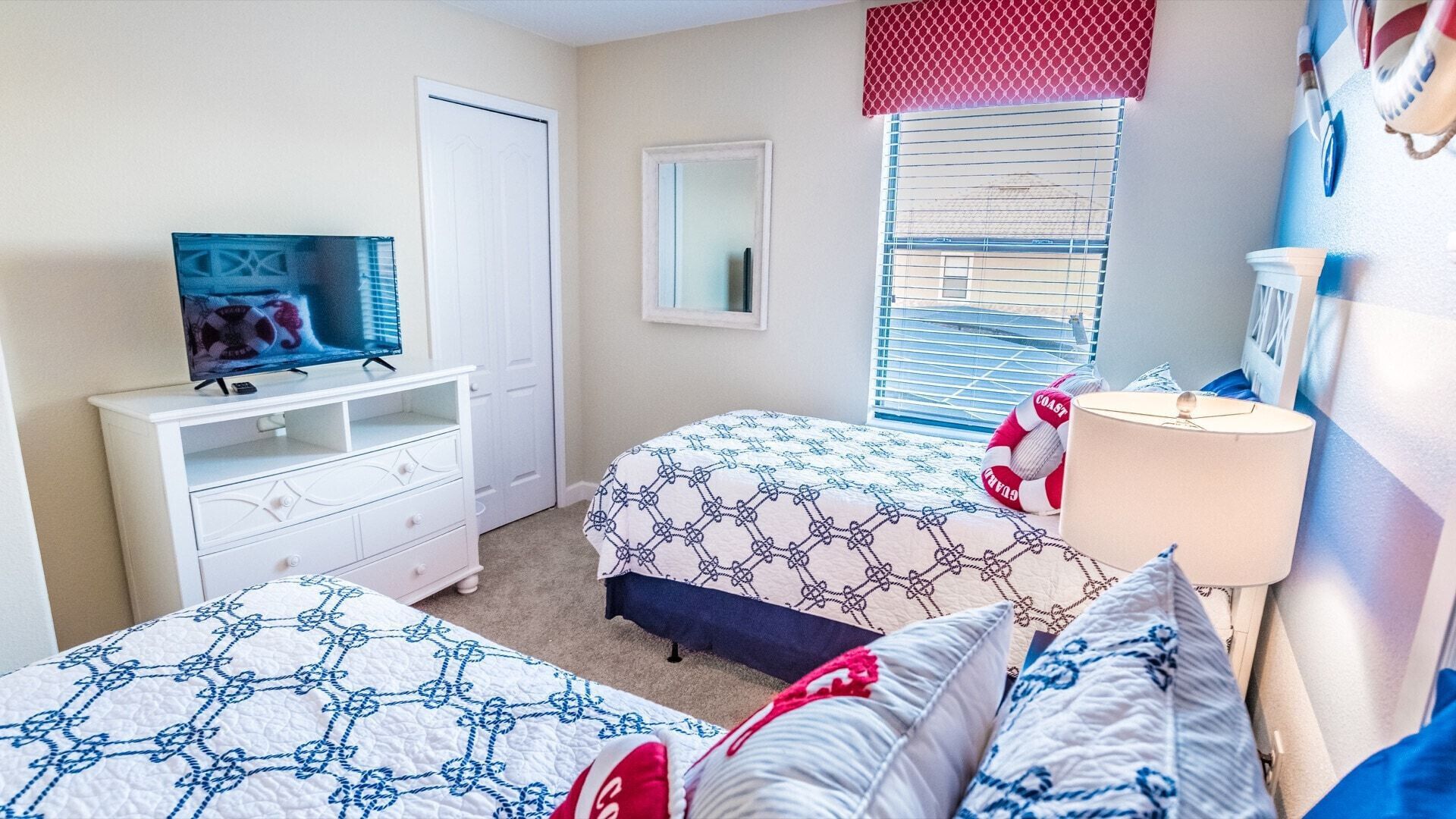 Une chambre avec deux lits, un téléviseur posé sur une commode, une fenêtre avec des stores et une lampe.