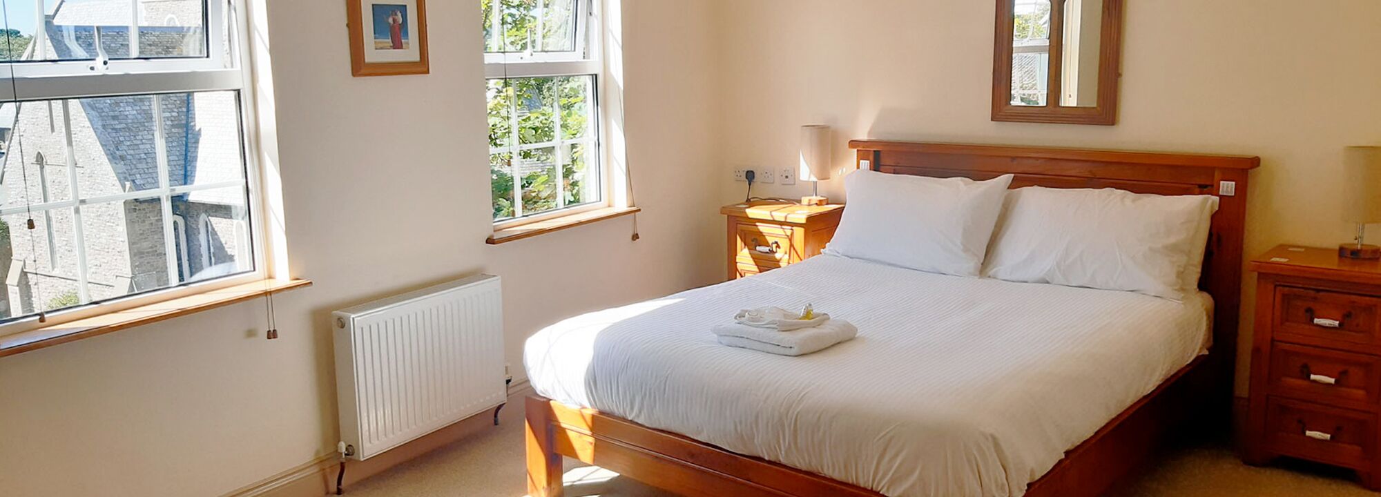 A neatly made bed with white linens, a bedside table with a lamp, and a window with a view of greenery.