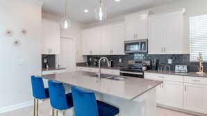 Uma cozinha moderna com armários brancos, uma ilha central com cadeiras azuis, eletrodomésticos de aço inoxidável e iluminação pendente.