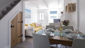 Zona pranzo con tavolo in legno apparecchiato per quattro persone, sedie bianche e vista su un soggiorno con divano e un quadro.