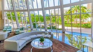 A modern hotel lobby with large windows, a curved sofa, and a round coffee table. - Beachfront  Luxury Resort | Views & Pools (Hallandale Beach)