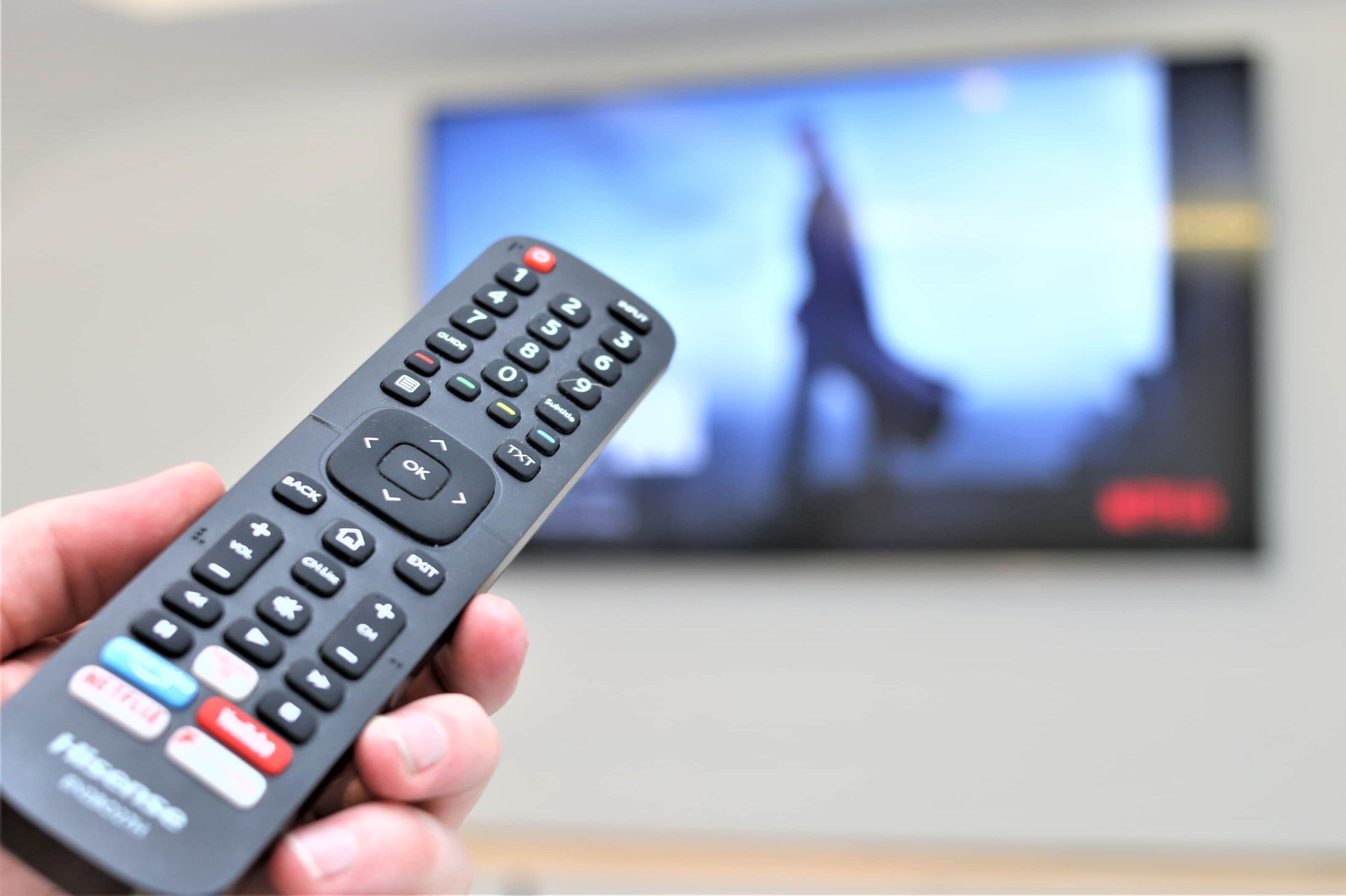 A hand holding a remote control in front of a television displaying a person.