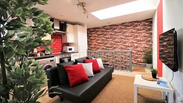 Un salon moderne avec un canapé noir, des coussins rouges et blancs, une petite table blanche et un téléviseur fixé au mur.
