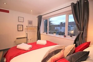 A hotel room with a bed, a window with curtains, a radiator, and a wall-mounted shelf with a plant.