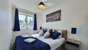 A bedroom with a bed, a nightstand, a lamp, a window with curtains, and a cityscape painting on the wall.