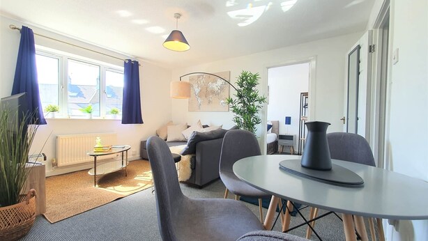 A modern living room with a sofa, chairs, a coffee table, and a dining table. There is a large window with curtains, a radiator, and a potted plant.
