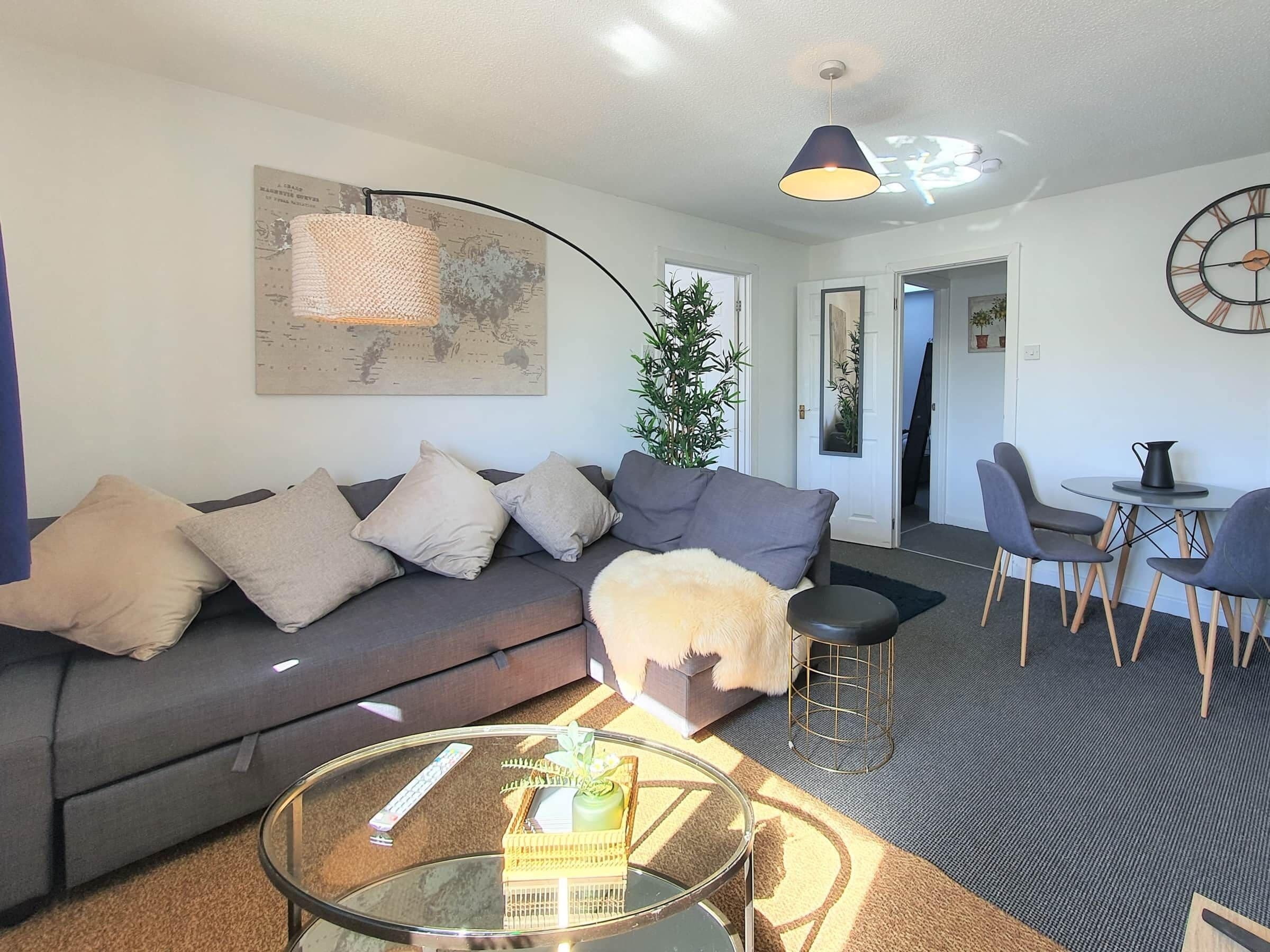 Een moderne woonkamer met een grijze bank, een glazen salontafel, een kleine ronde tafel met stoelen, een hanglamp en een wereldkaart aan de muur.