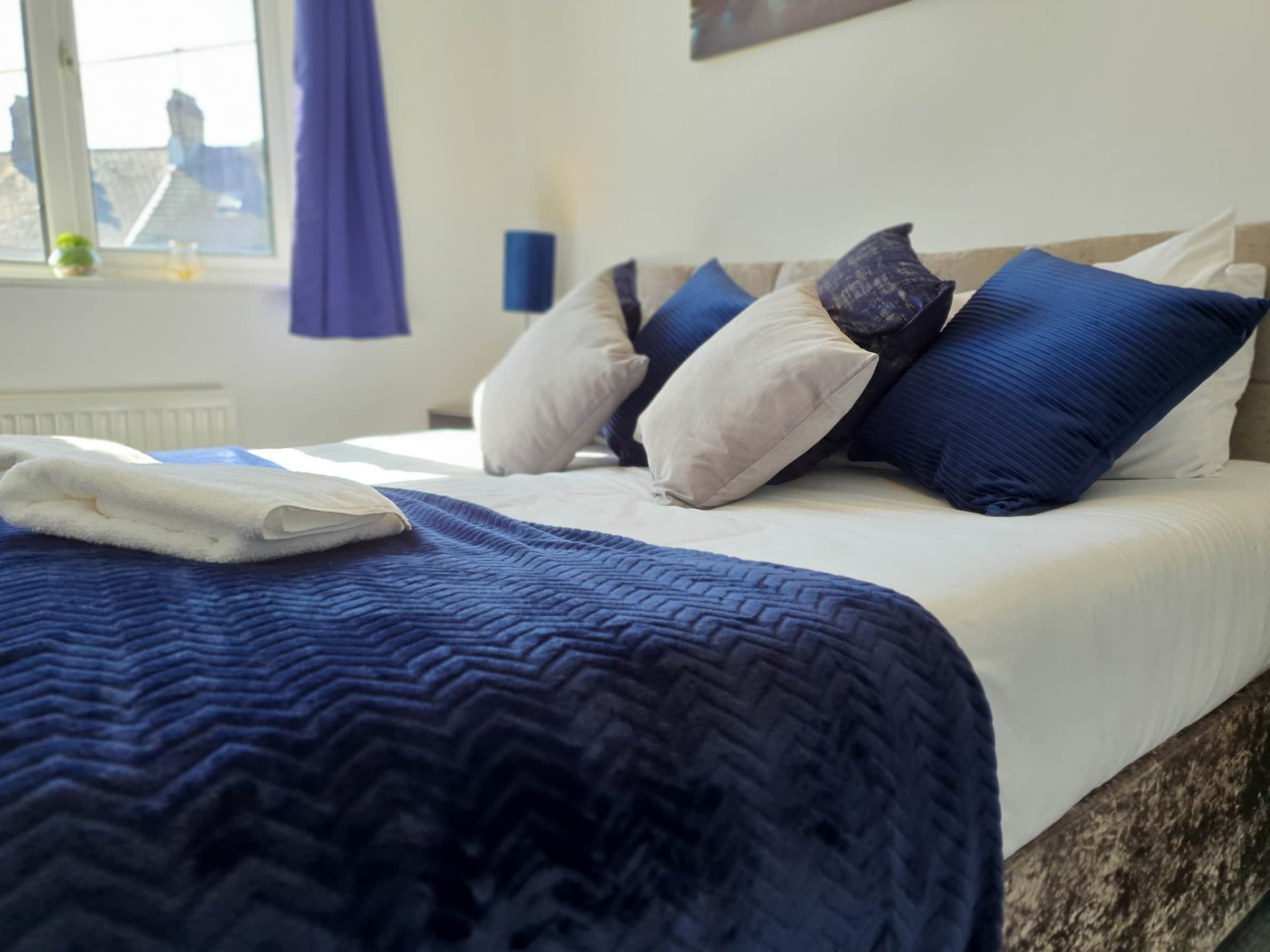 A neatly made bed with a blue quilt, multiple pillows in shades of blue and white, and a window with a view of buildings.