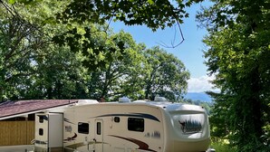 Una caravana grande estacionada en una zona boscosa con un porche cubierto y una vista panorámica de montañas al fondo.