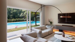 A modern living room with a large sliding glass door leading to a pool area, a flat-screen TV mounted on the wall, and a minimalist coffee table. - Villa Jaizubia Golf by FeelFree Rentals (Hondarribia)
