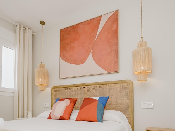 A bedroom with a bed, a headboard, a painting, two pendant lights, a window with curtains, and a wall socket.