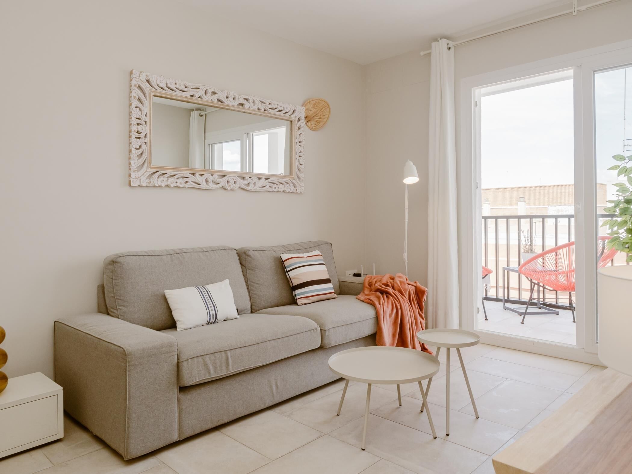 A modern living room with a sofa, two small tables, a mirror, and a balcony with red chairs.