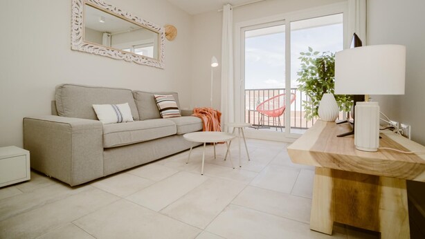 A modern living room with a grey sofa, a round table with a lamp, a large mirror, and a balcony with a chair.