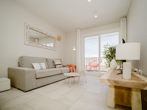 A modern living room with a grey sofa, a round table with a lamp, a large mirror, and a balcony with a chair.