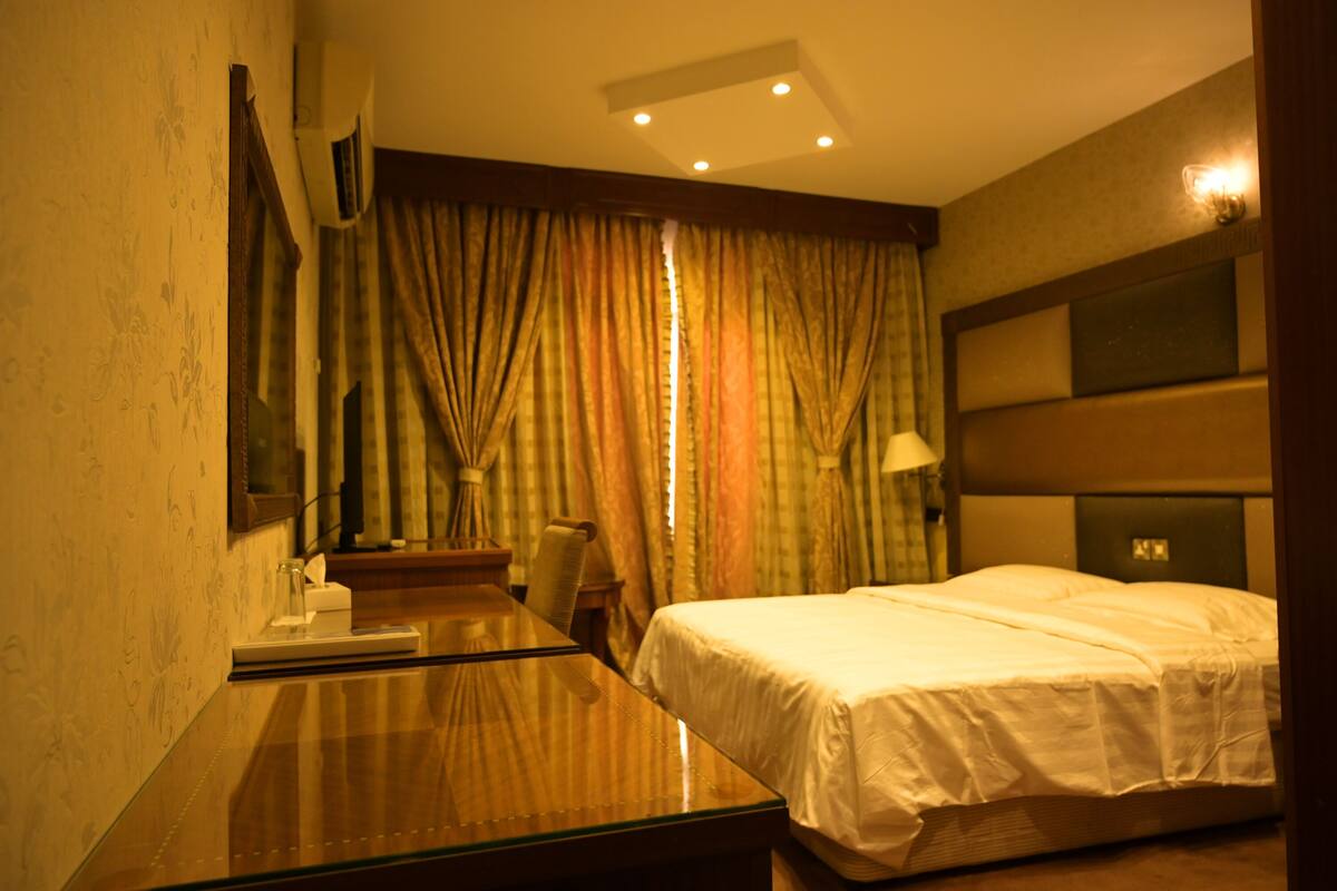 Ein Hotelzimmer mit einem Bett, einem Schreibtisch und einem Sessel. Es gibt ein Fenster mit Vorhängen, eine Deckenleuchte und eine Wand-Klimaanlage.