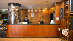 A reception desk with two staff members, a clock, framed pictures, and a sign indicating the reception area. - ABJAD CROWN HOTEL (Dubai)