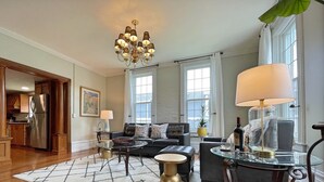 A spacious living room with a modern chandelier, a glass coffee table, a leather sofa, and a dining area with a glass table and chairs.