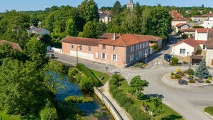 An aerial view of a town with a prominent red-roofed building, surrounded by lush greenery and a small body of water.