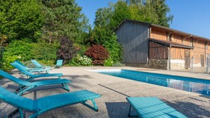 A swimming pool with lounge chairs, surrounded by trees and a building with a sloped roof.