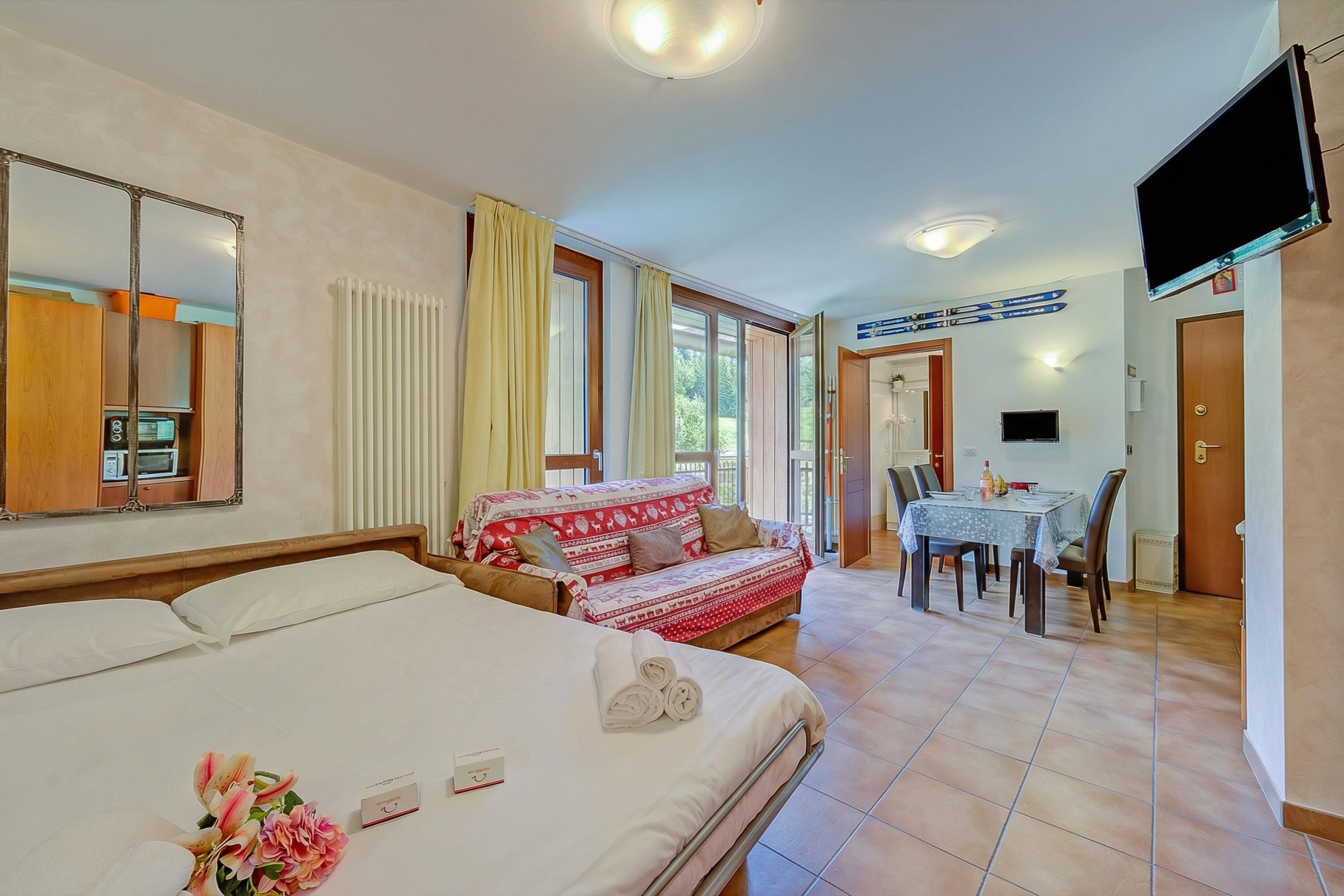 A hotel room with a bed, a sofa, a dining table with chairs, a TV, and a window with curtains.