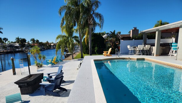 A swimming pool with lounge chairs and a covered area, surrounded by palm trees and a view of a marina. - Outdoor lovers dream home- Pool, outdoor kitchen, private dock, and so much more (New Port Richey)