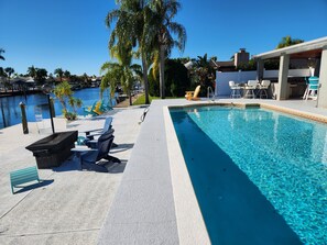 A swimming pool with lounge chairs and a covered area, surrounded by palm trees and a view of a marina. - Outdoor lovers dream home- Pool, outdoor kitchen, private dock, and so much more (New Port Richey)