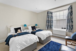 A bedroom with two beds, a window with curtains, a chair, and a bedside table with lamps.