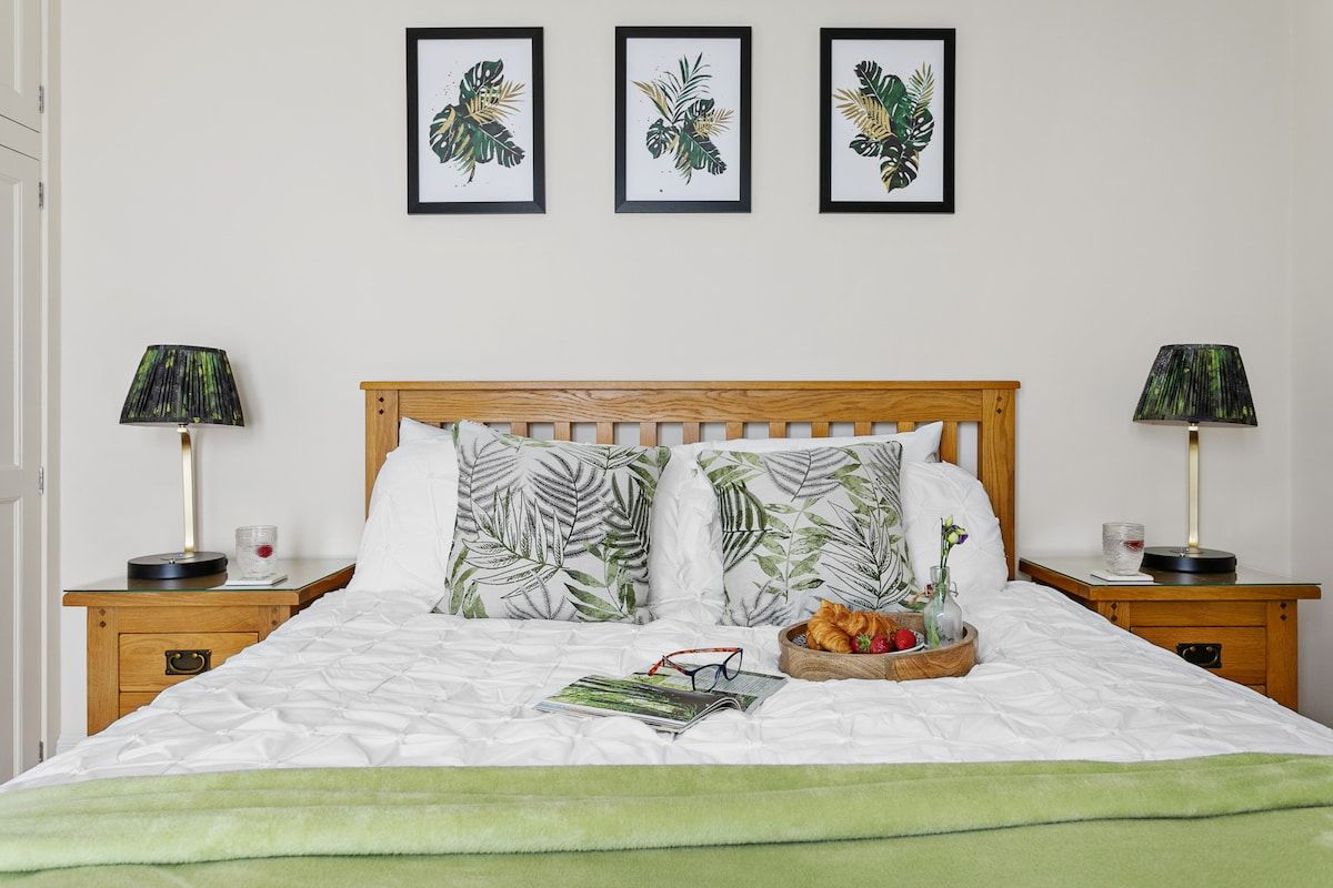 Una cama bien hecha con una colcha verde, dos almohadas con motivos de hojas, una mesita de noche con una lámpara y una bandeja con frutas y un jarrón.