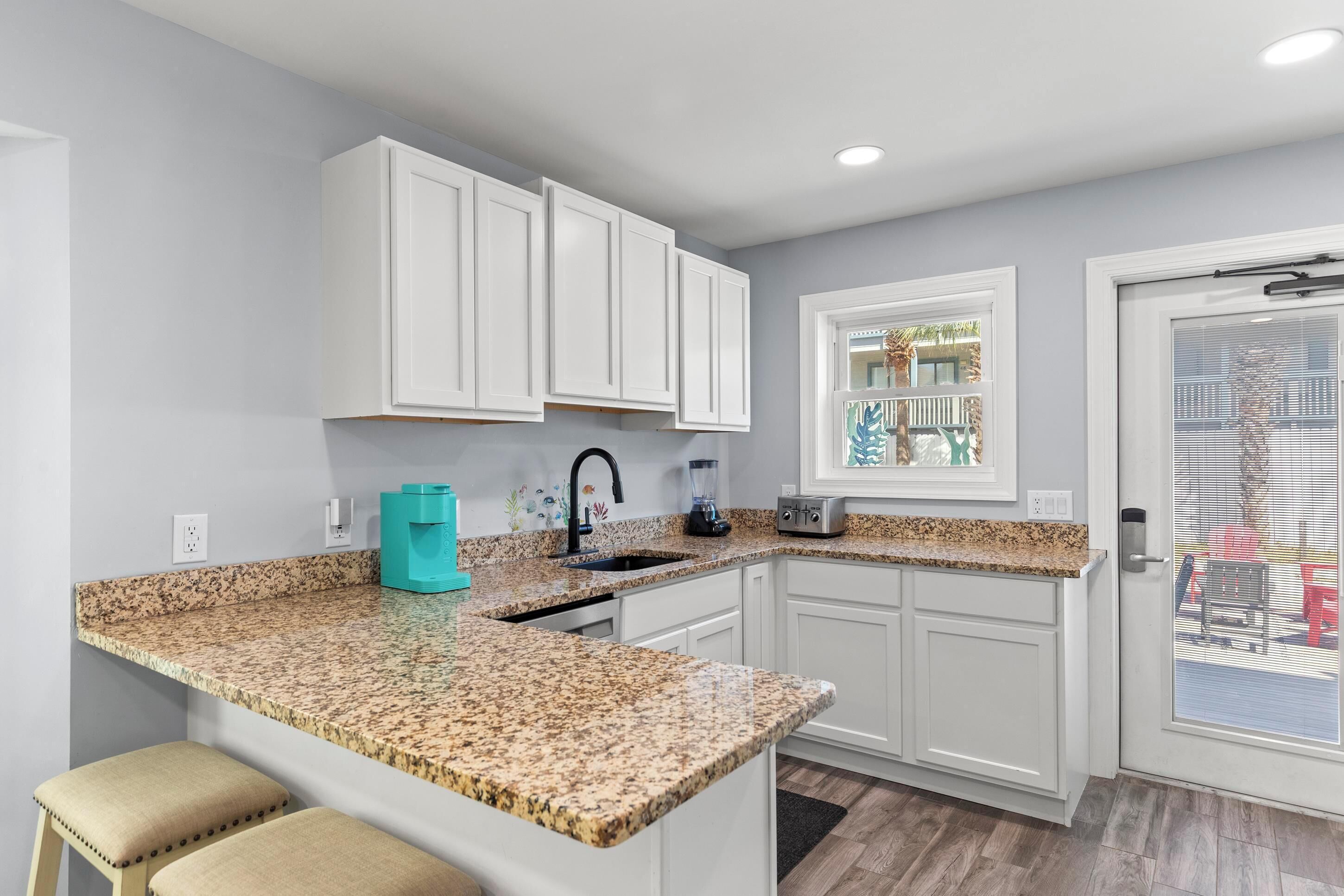 A kitchen with white cabinets, granite countertops, a coffee maker, and a window with a view of outdoor furniture.