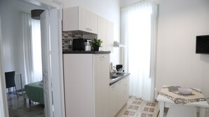 A compact kitchen with white cabinets, a small dining table with a chair, and a view of a bedroom with a green bedspread. - Palazzo Leonardo 1 (Scicli)
