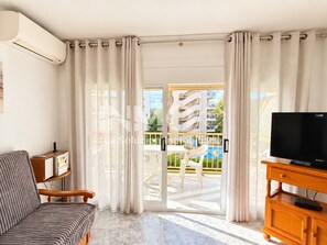 Un salon moderne avec un canapé, un téléviseur à écran plat posé sur un meuble en bois, et une porte-fenêtre coulissante donnant sur un espace extérieur avec une piscine et des chaises.
