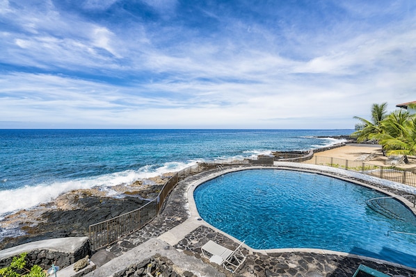 A circular pool with a stone border, adjacent to a rocky shoreline and a beach.