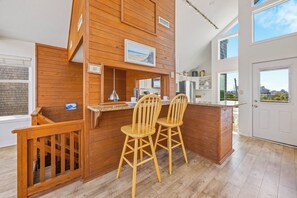 A modern kitchen with wooden accents, a bar area with two stools, and a view of the outside. - Stunning Oceanfront Home with Hot Tub, Grill, and Stellar Ocean Views (Avon)