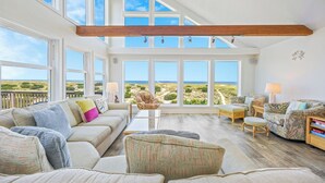 Ein geräumiges Wohnzimmer mit hoher Decke, großen Fenstern und Blick auf den Strand.