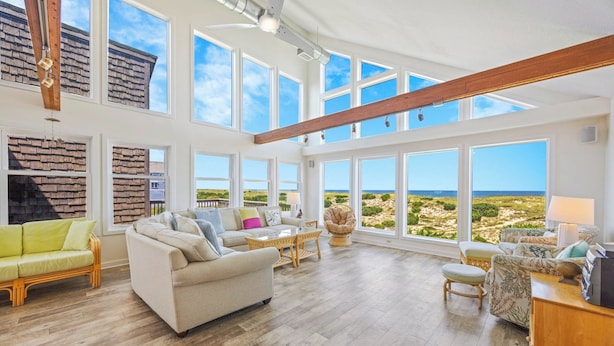 Ein geräumiges Wohnzimmer mit großen Fenstern, einer Holzbalkendecke und Blick auf den Strand.
