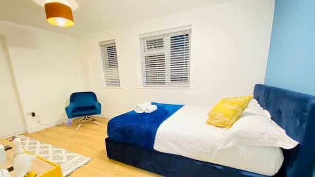 Une chambre avec un lit bleu, des oreillers blancs et un coussin jaune. On y trouve une chaise bleue et une petite table sur laquelle se trouve un objet jaune.