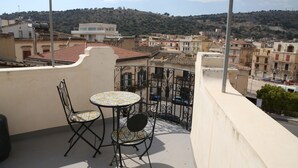 A balcony with a table and two chairs, overlooking a cityscape with buildings and a hill in the background.