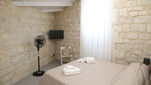 A room with stone walls, a bed with a beige bedspread, a standing fan, a chair with a decorative pillow, a flat-screen TV mounted on the wall, and a window with white curtains.