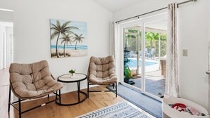 Ein Zimmer mit zwei gepolsterten Sesseln, einem kleinen Tisch, einem Strandbild und einer Glasschiebetür, die zu einem Außenpoolbereich führt.